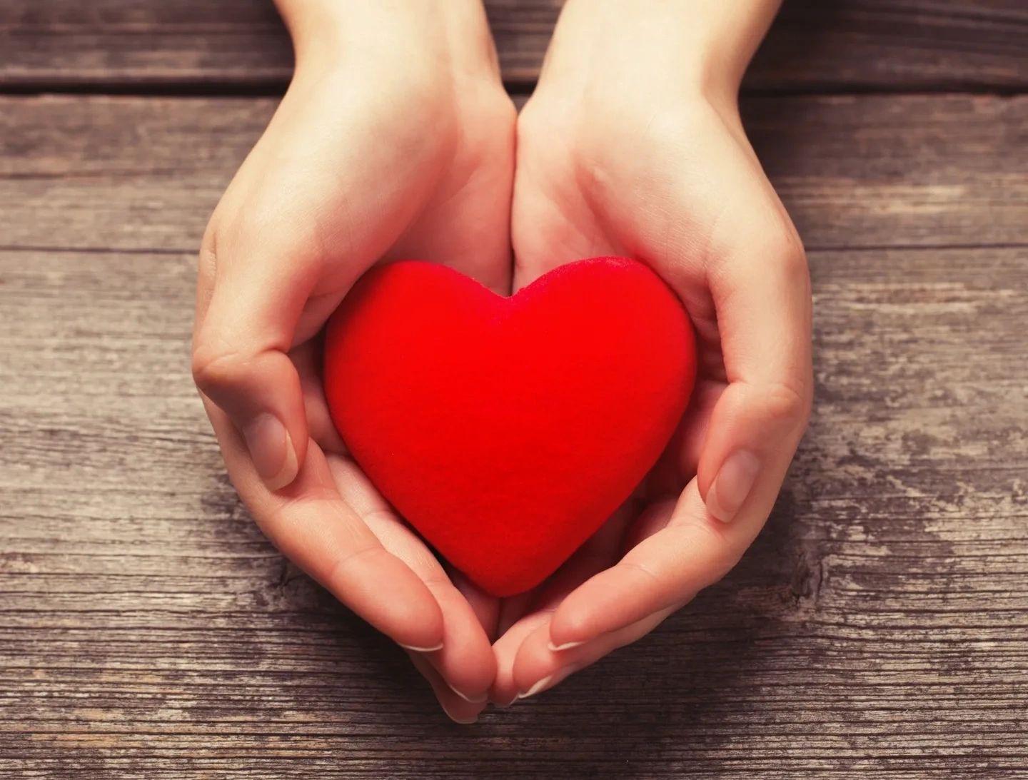 Hands holding a red heart on wooden surface.