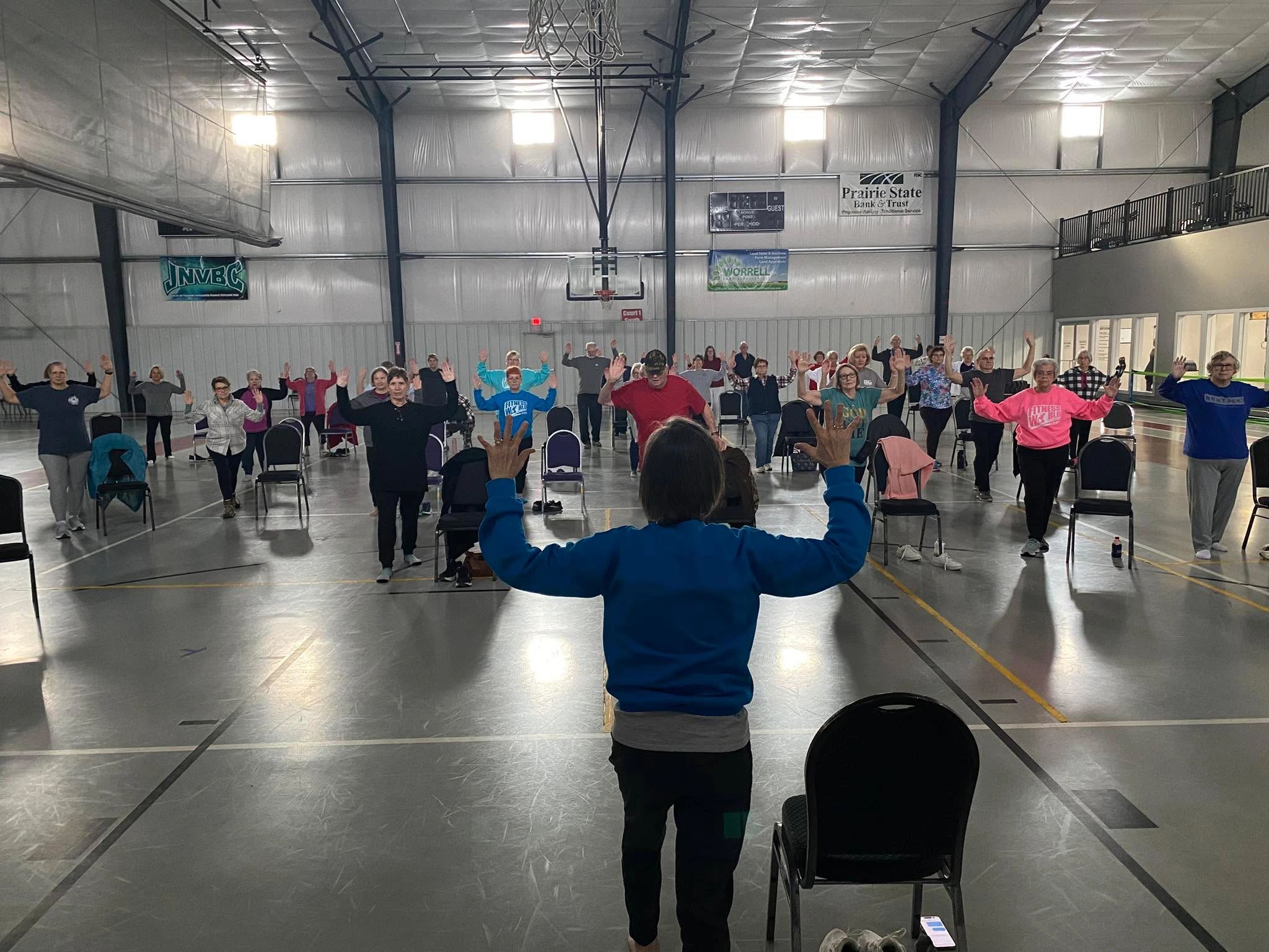 Group fitness class in a gym with instructor and participants raising hands.