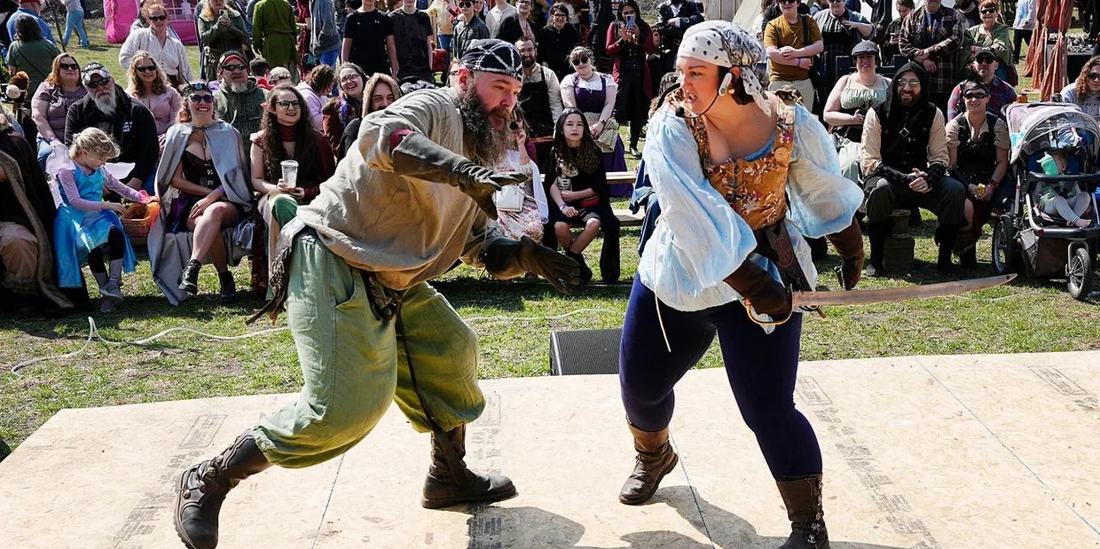 Two people engage in a staged sword fight at a lively outdoor festival with an audience watching.