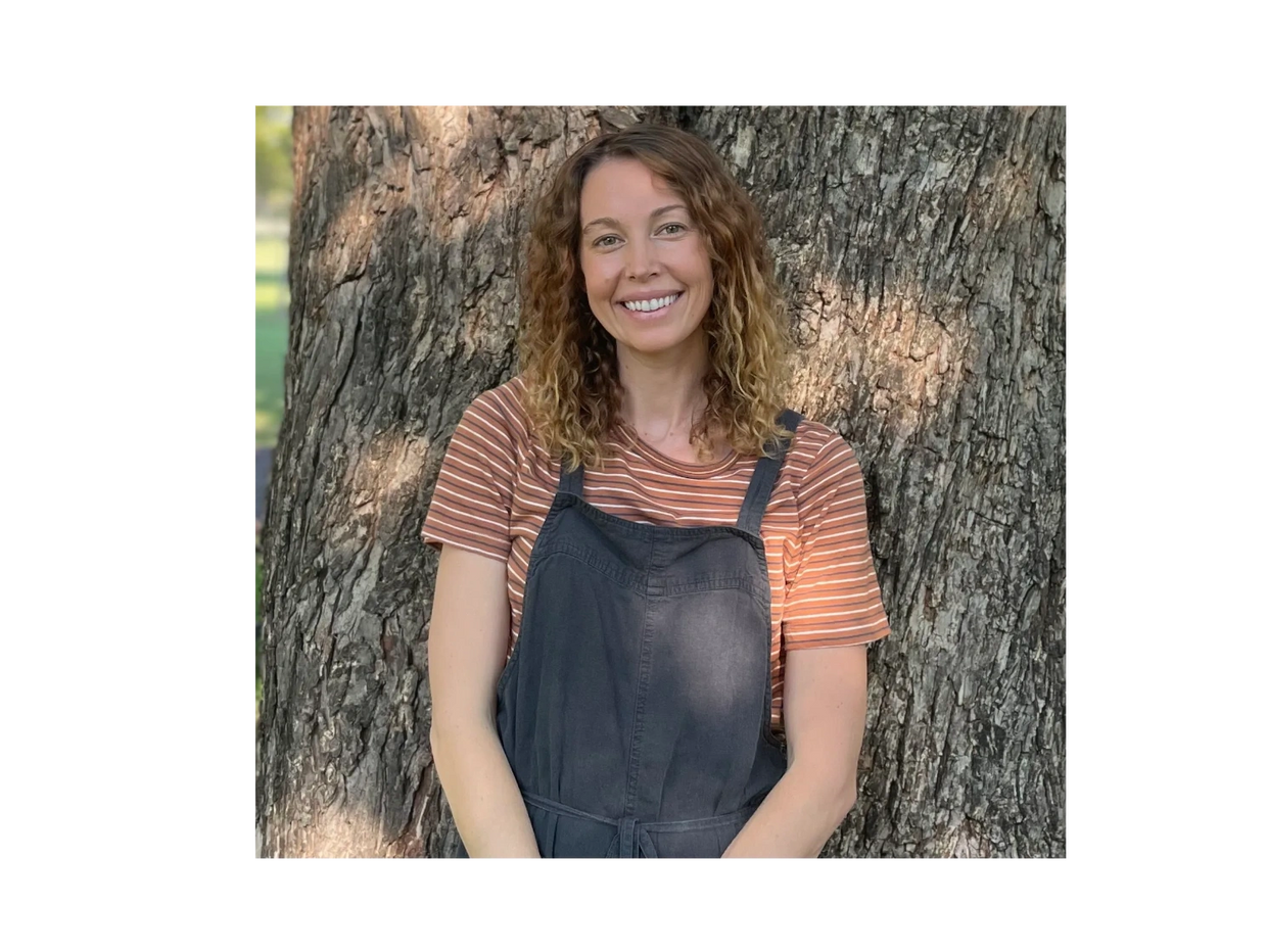 Smiling woman with curly hair stands in front of a large tree.