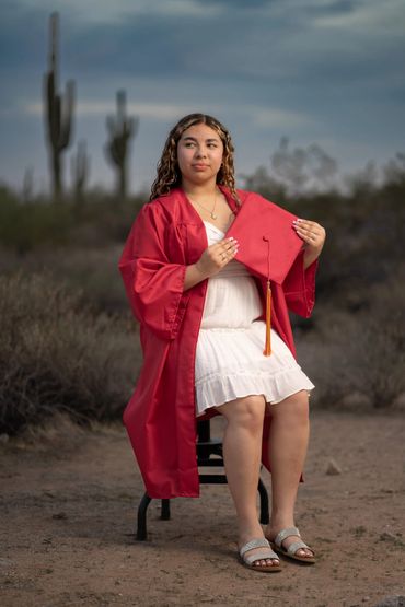 Young woman in a red graduation gown holding a cap outdoors in a desert setting.