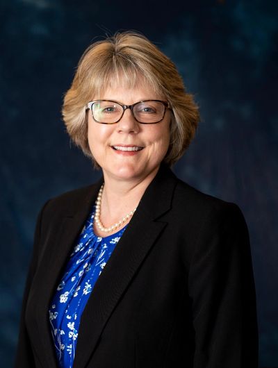 Professional woman in glasses and black blazer smiling against a dark blue background.