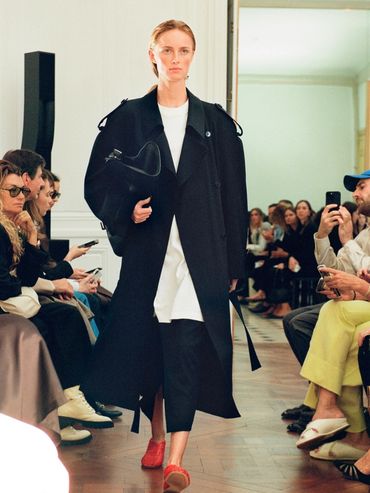 Model walks runway in oversized black coat, white top, black pants, and red shoes at fashion show.