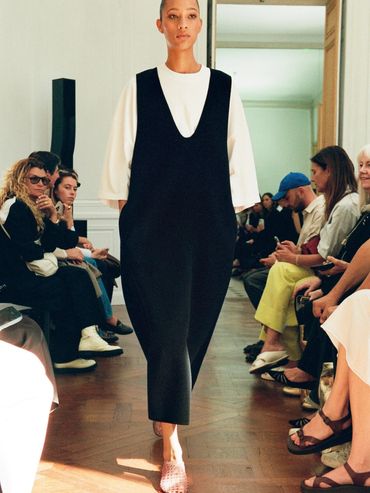 Model walking down runway in a minimalist black dress over a white top.