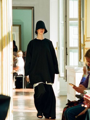 Model wearing oversized black outfit and hat on runway in elegant room.