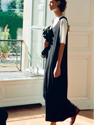 Woman in a black dress and white shirt standing near a sunlit window.