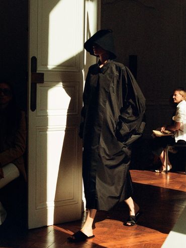 Model in oversized black coat and wide-brim hat walking indoors with dramatic lighting.