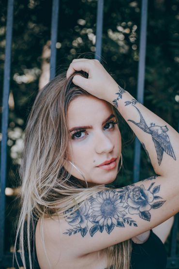 Young woman with floral and bird tattoos poses confidently outdoors.