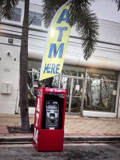 Red ATM machine with a yellow "ATM HERE" flag by a palm tree.