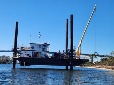 A workboat lifted above water by four large vertical legs near a shore with a crane in the background.