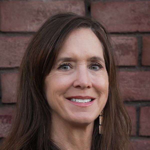 Smiling woman with long brown hair in front of a brick wall.