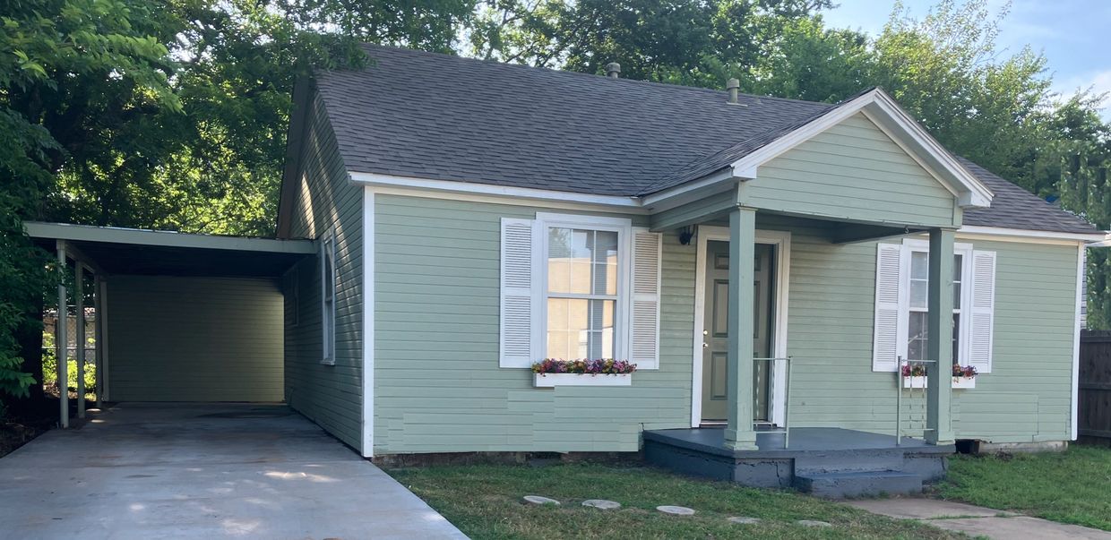 Small green house with white shutters and a carport on the side.