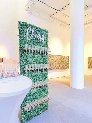 A green wall with "Cheers" sign and champagne glasses on shelves in a bright room.