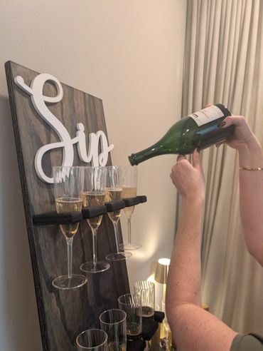 Person pouring champagne into glasses on a wooden display with 'Sip' sign.