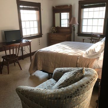 Fully furnished guest room with  antique Victorian dresser, plush inner springs mattress, smart TV, ceiling fan, classic trim, padded low pile carpet, swing arm lamp, two wooden Cushman chairs, swivel rocking upholstered bedroom chair.