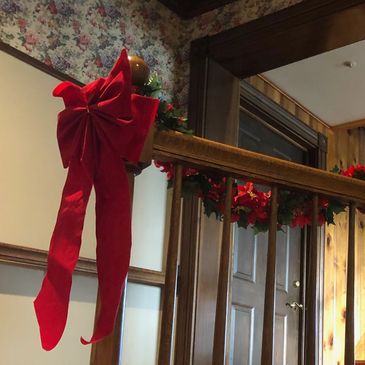 Oil rubbed finished banister decorated with red velvet ribbons, greenery and poinsettias. and oak handrail ADA interior ramp from the dining room to the sitting room, floral wallpaper header, dark stained custom millwork casing and rosettes.