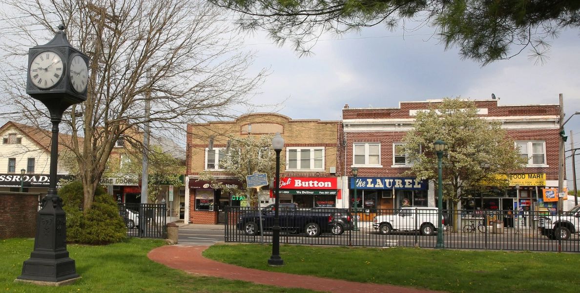 A small town street with shops, a clock tower, and trees in a park.