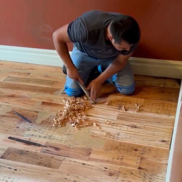Man kneeling scraping a wooden floor with wood shavings scattered around.