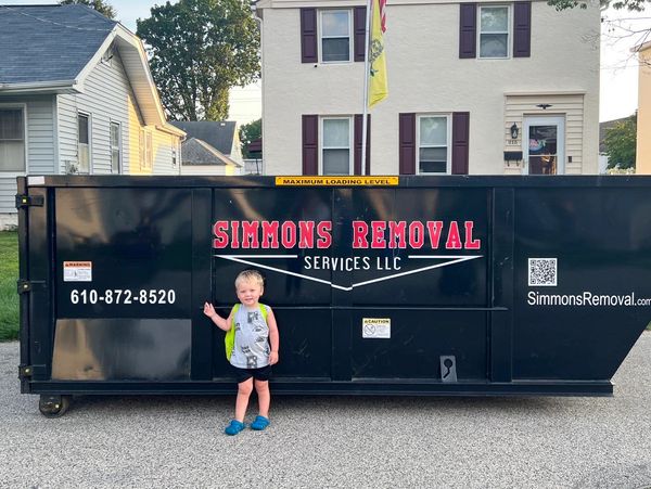 A toddler stands in front of a large Simmons Removal dumpster on a residential street.