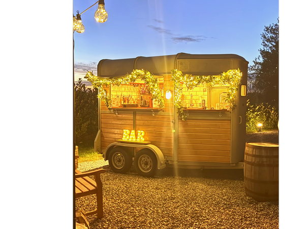 A cozy outdoor bar setup in a trailer decorated with string lights and greenery at dusk.