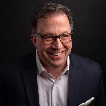 Smiling man wearing glasses, a dark blazer, and a white shirt against a black background.