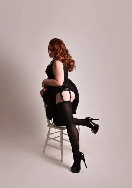 A woman with red hair, a black dress, and a garter belt kneeling on a chair in high heels 
