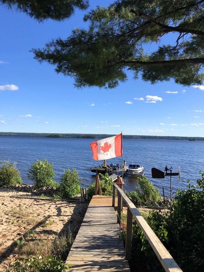 A wooden dock with a Canadian flag overlooking a calm lake under a clear blue sky.