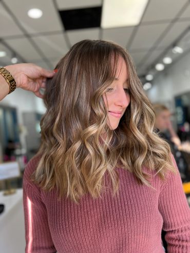 Woman with wavy, highlighted hair wearing a pink sweater in a salon.