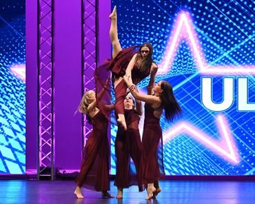 Four dancers in maroon dresses perform a lift on stage with a bright star backdrop.