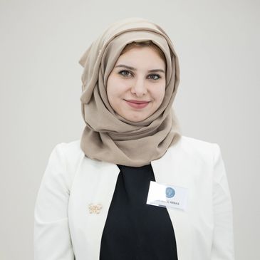Young woman wearing a beige hijab and white blazer smiling at the camera.