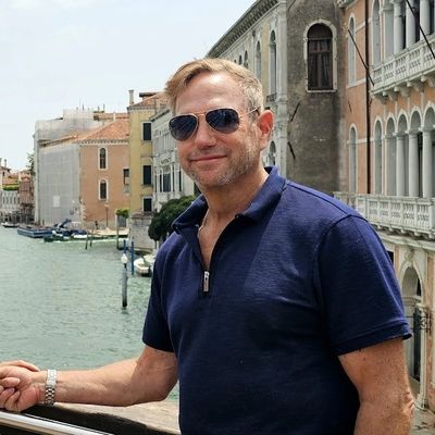 Man in sunglasses standing by a canal with historic buildings in the background.