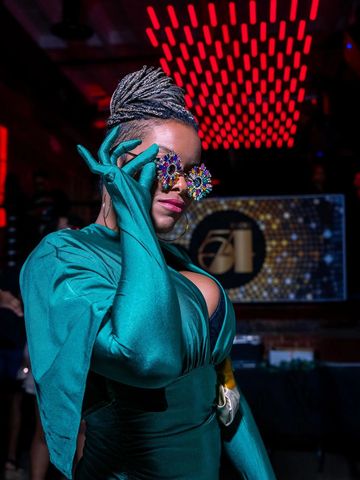Person in teal outfit and colorful glasses posing confidently in a stylish, dimly lit venue.