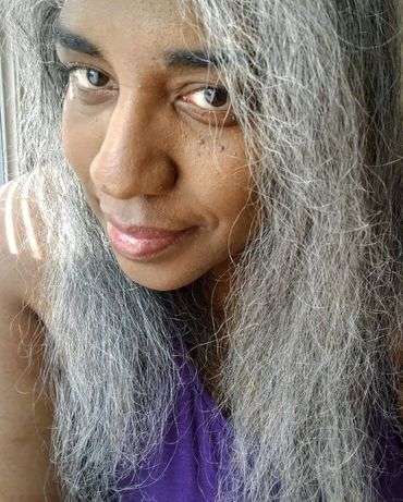 Image of a smiling gray haired Black Latine femme wearing a sleeveless purple top. Three streaks of