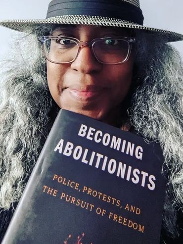 Image of the essayist a Black Latine femme wearing a Panama Straw Hat holding a copy of a book