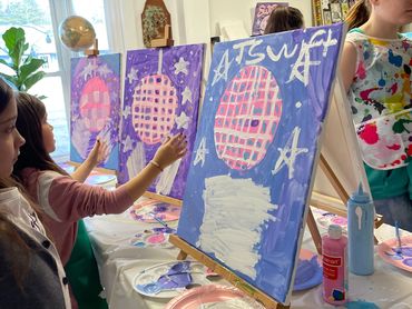 Children painting pink disco balls on purple canvases in an art class.
