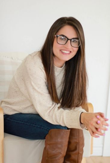Smiling woman with glasses sitting, wearing sweater and brown boots.