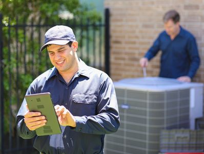 Smiling HVAC technician in a cap using a tablet with a colleague working on an outdoor AC unit