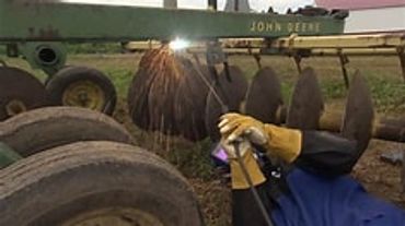 Person welding a disc harrow under a John Deere frame outdoors.