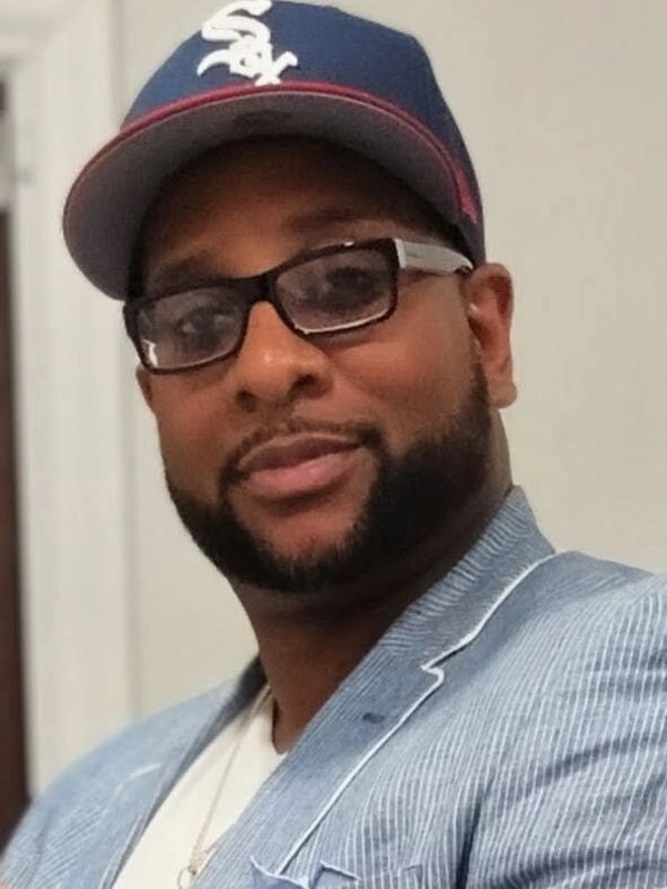 Man wearing glasses, a blue and white cap, and a pinstripe blazer, smiling slightly.