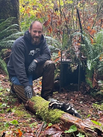 Man in outdoor gear kneeling near a skunk in a forest.