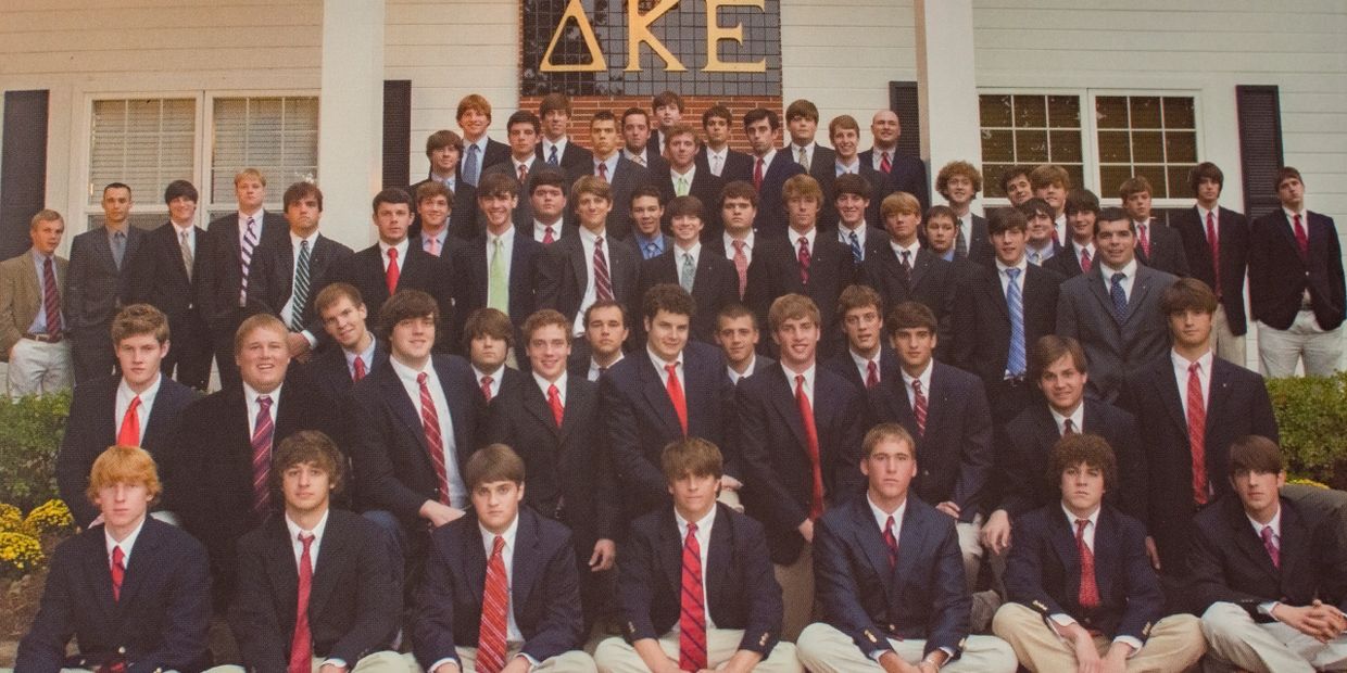 Group photo of a fraternity in suits and ties outside a house with Greek letters.