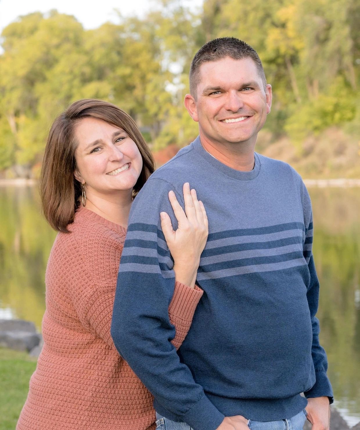 Smiling couple posing outdoors by a lake with fall foliage.