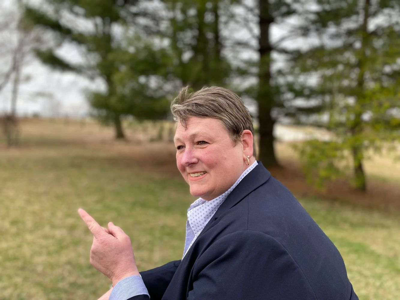 Smiling person pointing outdoors with greenery in the background.
