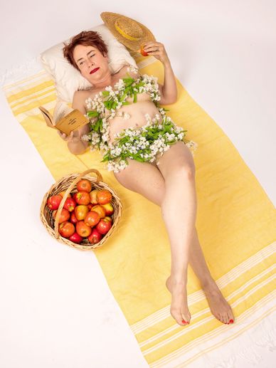 Woman lying on yellow towel wearing flowers, holding a book and straw hat with basket of apples nearby.