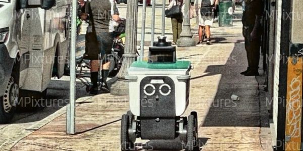 Delivery robot navigating a busy urban sidewalk with pedestrians and vehicles.