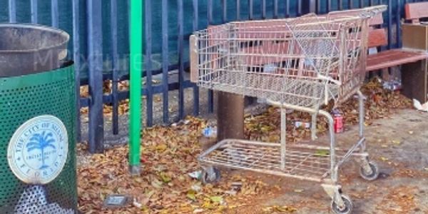 Empty shopping cart next to a park bench and trash can outdoors.