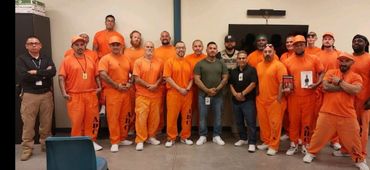 Group of men, some in orange prison uniforms, posing indoors against a plain wall.