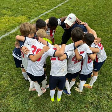 Youth soccer team huddling with coach on grassy field.