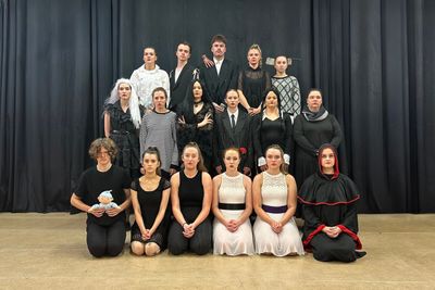A theatrical group poses in costume on stage against a black curtain backdrop.