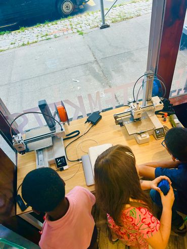 Three kids watch and work with two 3D printers on a wooden table near a window.